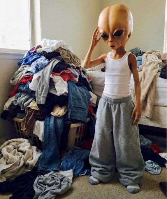 An alien figure stands in a bedroom surrounded by massive piles of unfolded laundry, wearing a tank top and sweatpants while holding its head in apparent frustration. The image humorously depicts the overwhelming dread of tackling laundry, matching the "gm ct" (good morning crypto Twitter) sentiment with a relatable morning struggle.
