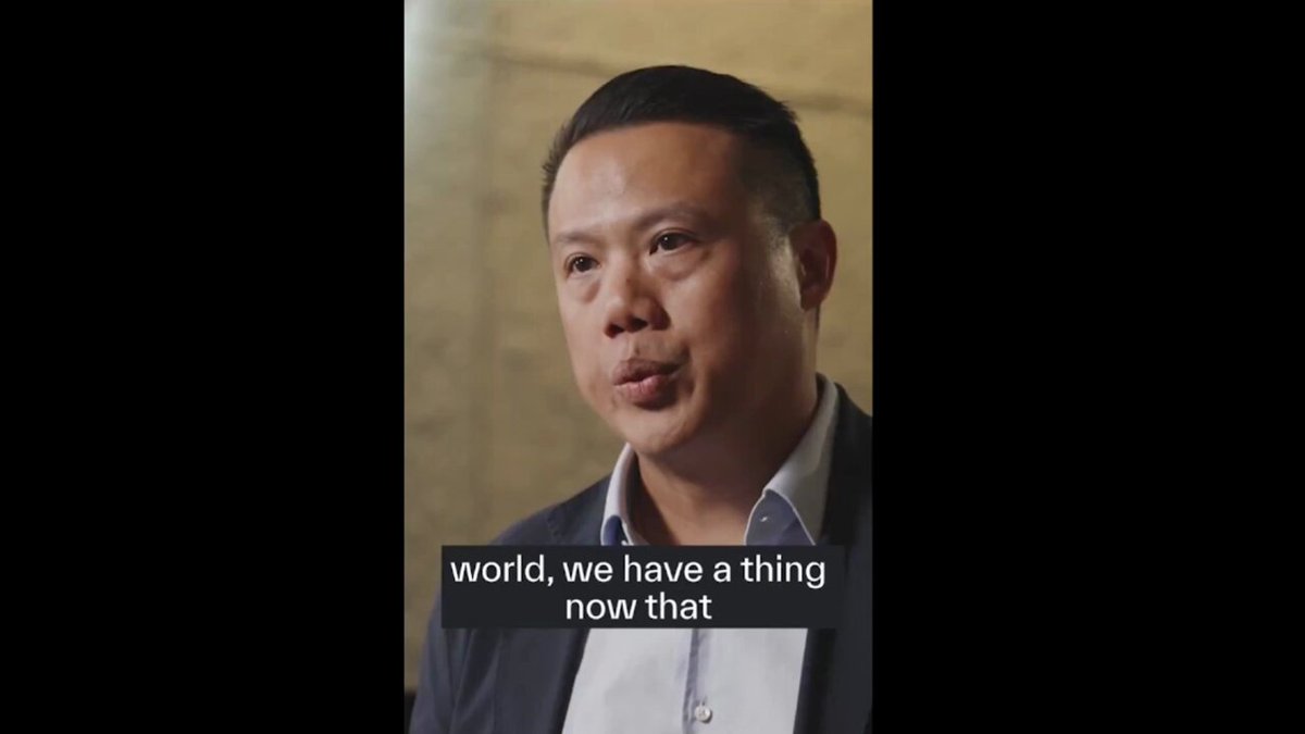 The image features a man wearing a suit, standing against a gray background with white text that reads "world, we have a thing now that". The man appears to be talking about something related to blockchain technology or cryptocurrency, as indicated by the accompanying post on social media.
