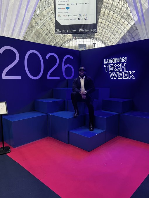 The image features a man sitting on top of blue boxes, posing for a photo at an event called "London Tech Week" with a sign that reads "2026". The man appears to be wearing a suit and tie, indicating a formal occasion or gathering. There is also a TV screen in the background displaying information about the event.