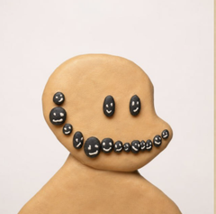 A pale clay figure with a smooth, featureless face except for a grin made of black, shiny beads or stones.