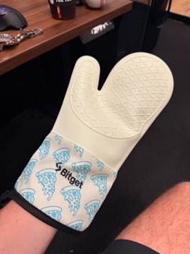 A foot clad in a cream-colored "Spitget" branded oven mitt with a quilted top and pizza-patterned cuff, humorously illustrating the comparison between high-end market-making perks and the modest branded merchandise received in return.