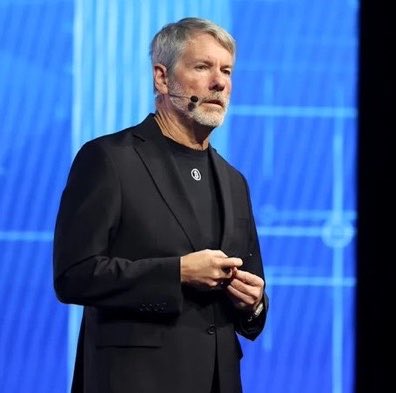 A bearded man in a black suit with a small circular pin stands on stage against a blue backdrop, wearing a headset microphone as he addresses an audience. The image relates to the post about Strategy's recent purchase of 4,603 bitcoin, likely depicting Michael Saylor, the executive chairman known for championing corporate bitcoin adoption.