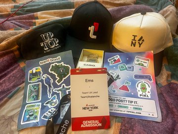 An assortment of merchandise is laid out on a bedspread, including two baseball caps (black and cream), a black "I <3 NY" beanie, sticker sheets, a "Coach C414043" name tag, and a lanyard reading "TeamUKLMG TeamRainbowswfc GENERAL ADMISSION" and "FOOD POST TIP 11".