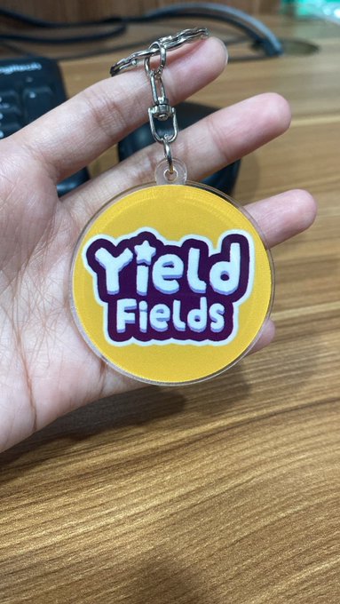 A hand displays a circular acrylic keychain featuring the "Yield Fields" logo in white text on a yellow background with purple accents, attached to a silver chain—showcasing branded merchandise for the Yield Fields project.