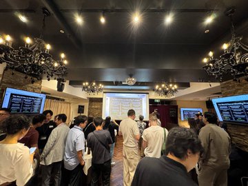 A crowded indoor event or presentation where a group of people stand watching a slide displayed on a screen, with chandeliers hanging from a dark ceiling.