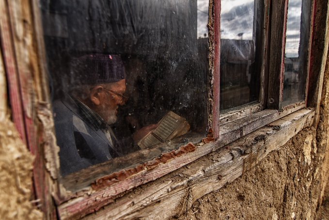 An older man wearing glasses and a traditional cap reads from a book behind a weathered, dirty window frame.