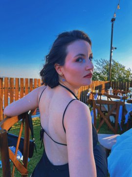 A photograph of a woman with dark hair in an updo, wearing a black backless dress, looking over her shoulder at the camera in an outdoor setting with wooden fencing.