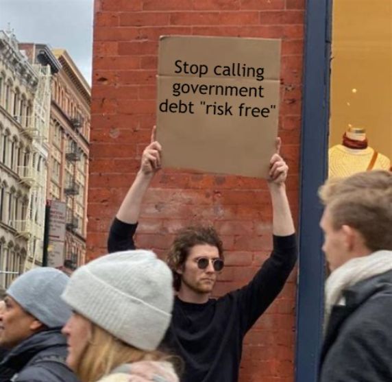 A man in sunglasses holds up a protest sign reading 'Stop calling government debt "risk free"' on a city street.
