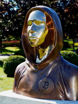 A bronze statue of a hooded figure wearing sunglasses stands outdoors, featuring a Bitcoin symbol (₿) prominently displayed on its chest. The artwork serves as a tribute to Bitcoin's mysterious creator, Satoshi Nakamoto, visually embodying the post's message about Bitcoin's pseudonymous founder and the ideological divide between believers and skeptics in the crypto community.