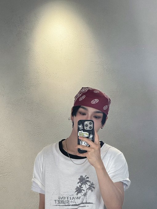 A mirror selfie of a young man wearing a maroon paisley bandana, a white graphic t-shirt, and a silver chain, holding a phone with a black sticker-covered case.
