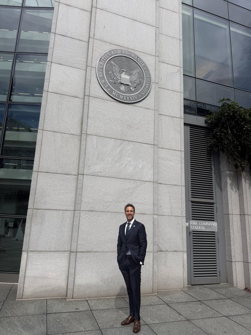 A man in a dark suit stands smiling in front of the U.S. Securities and Exchange Commission (SEC) building, with the agency's official seal visible on the stone facade.