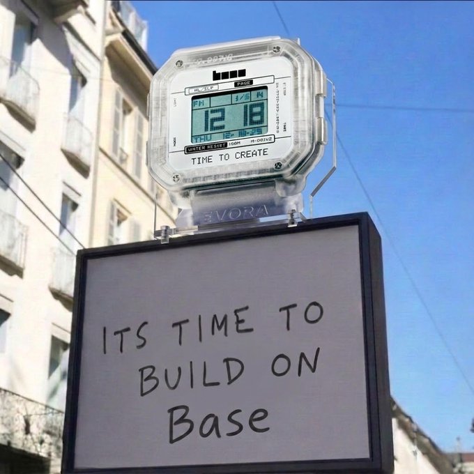 A large white wall clock by VORA mounted on a building displaying '12:18 PM THU 1/18/26' with a sign below reading 'ITS TIME TO BUILD ON Base' in handwritten-style text.