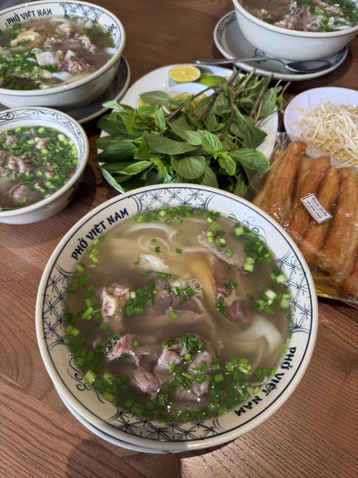 A bowl of "Phở Việt Nam" filled with beef, noodles, and chopped scallions sits on a wooden table alongside fresh herbs, bean sprouts, fried dough sticks, and additional bowls of the same soup. The image illustrates a Vietnamese dining experience, though the post focuses on using Binance to exchange stablecoins for local currency while traveling in Vietnam.