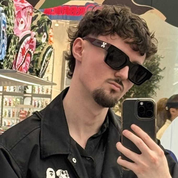 A mirror selfie of a curly-haired man wearing sunglasses and a black jacket, standing inside a brightly decorated retail store.