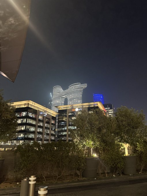 A nighttime cityscape featuring modern illuminated skyscrapers in the background with potted plants and trees in the foreground.