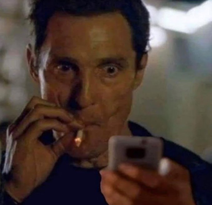A wide-eyed man smokes a cigarette while intently looking at a white smartphone screen.