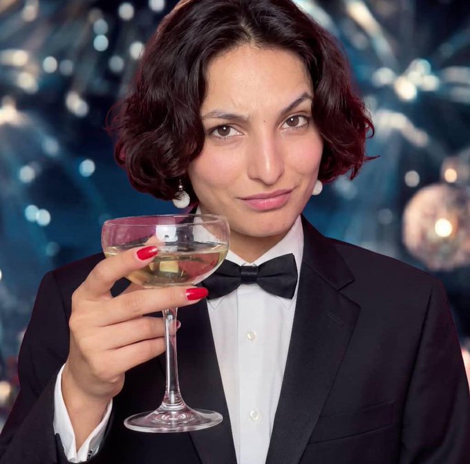 The image features a woman wearing a black suit, holding a glass of wine with a smile on her face. The background includes a blurred party scene with lights and people, suggesting that she might be at a social event or celebration. The caption reads "Elon don pay Make I go book that baddie for the weekend I'M ACTUALLY supporting my mutuals wbt you?". This implies that the woman is enjoying herself and possibly discussing her thoughts on the book with others present in the party scene.