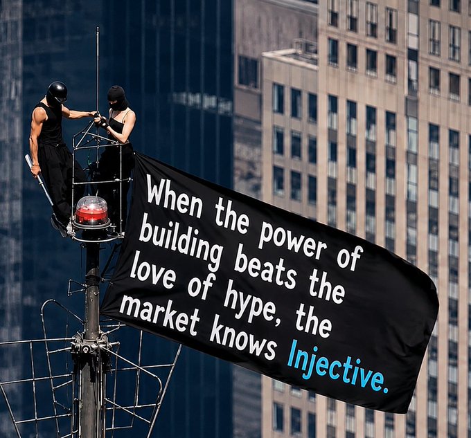 Two workers in black masks hang a black flag from a rooftop pole against a city skyline, displaying the text: 'When the power of building beats the love of hype, the market knows Injective.'