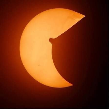 An orange-hued partial solar eclipse with the moon covering the right portion of the sun, showing a clear crescent shape against a dark background.
