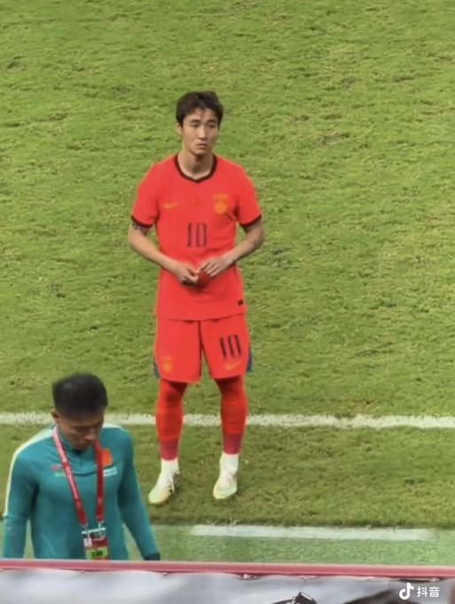 A soccer player wearing a red number 10 jersey and shorts stands on a grass pitch, with a Chinese social media watermark in the corner.