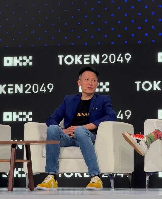Binance CEO Richard Teng sits on stage at TOKEN2049, wearing a "BINANCE" branded t-shirt under a blue blazer with yellow sneakers, as he discusses the growing demand for tokenized stocks and private market products amid information flow challenges.