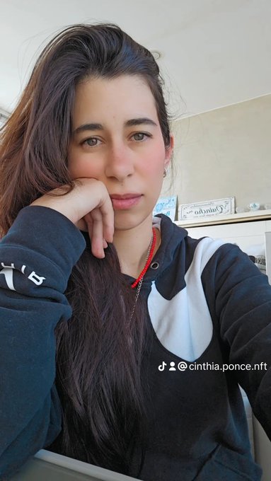 A selfie of a woman with long dark hair resting her chin on her hand, wearing a black and white hoodie and a red beaded necklace, with a TikTok handle '@cinthia.ponce.nft' visible.
