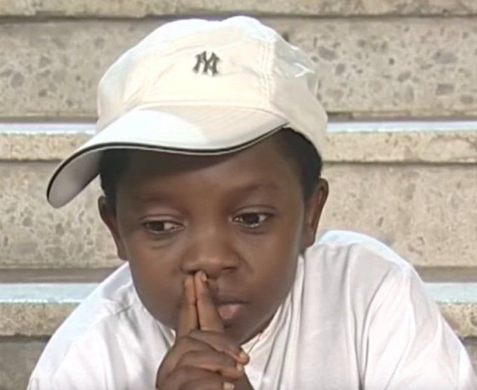 A young boy wearing a white New York Yankees cap, looking thoughtful with his finger near his face.