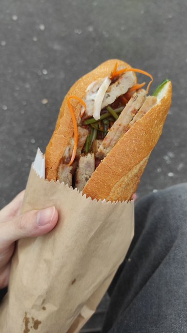 A hand holds a Vietnamese bánh mì sandwich wrapped in brown paper, filled with sliced meat, pickled carrots, daikon, and cucumber. The image's casual, on-the-go vibe contrasts with the post's discussion of "FDV" (Fully Diluted Valuation) in the crypto market, humorously comparing the thin crust of the sandwich to the fragile boundaries between real value and inflated market caps.