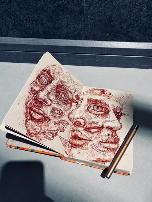An open notebook displays intricate red ink sketches of distorted, surreal faces with multiple eyes, accompanied by a metallic pen resting on its colorful cover.