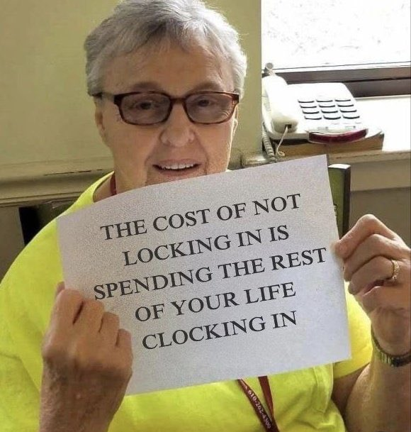 An older woman with short gray hair and glasses smiling while holding a sign that reads 'THE COST OF NOT LOCKING IN IS SPENDING THE REST OF YOUR LIFE CLOCKING IN.'