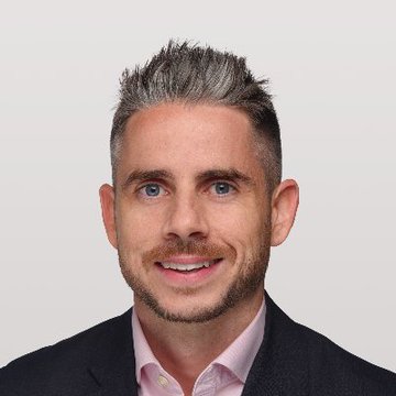 A professional headshot of a man with styled gray-tipped dark hair, a short beard, and a pink collared shirt under a dark blazer, smiling at the camera—likely a portrait of Ripple executive Reece Merrick referenced in the post about crypto payments.