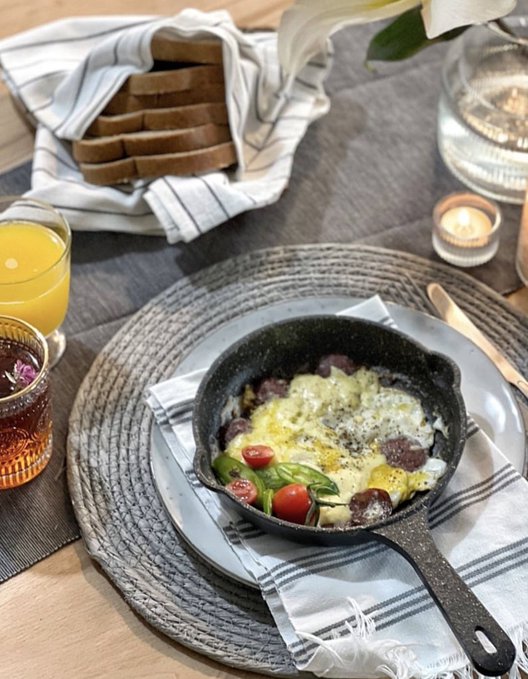 A breakfast scene featuring a cast-iron skillet with eggs, tomatoes, and sausage, placed on a striped napkin over a woven placemat, alongside a glass of orange juice, a basket of bread, and a lit candle.