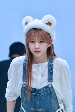 A young woman wearing a white furry bear-ear headband and a denim overall dress over a white shirt, posing for a portrait.