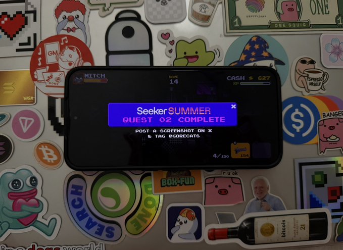 A smartphone resting on a surface covered in stickers displays a game screen reading 'SeekerSUMMER QUEST 02 COMPLETE POST A SCREENSHOT ON X & TAG @GORECATS' with CASH $627 and 14 MINS remaining.