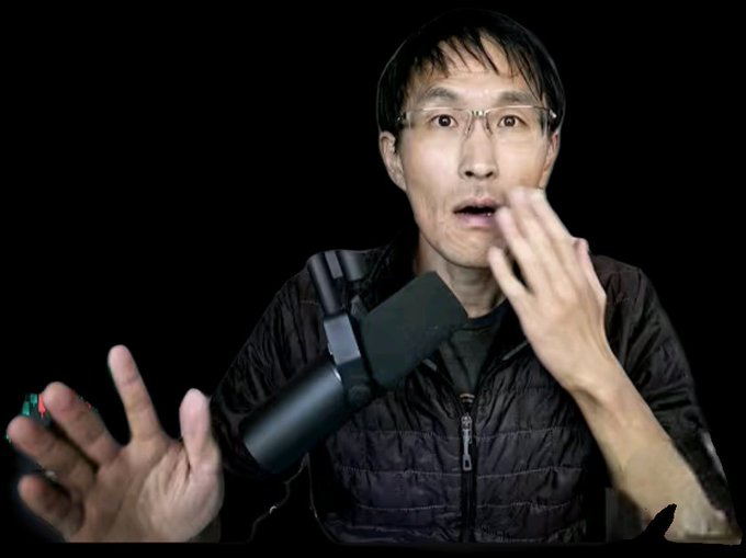 An Asian man with glasses and a black jacket gestures expressively while speaking into a microphone, holding a small object in his other hand against a black background.