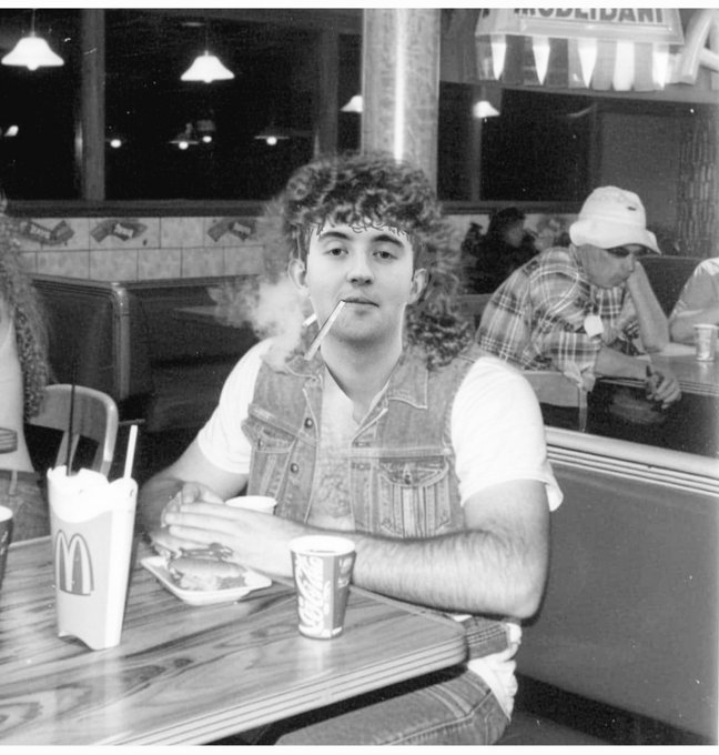 A black-and-white vintage photograph of a young man with curly hair and a bandana smoking a cigarette at a McDonald's, with a Coca-Cola cup and McDonald's fries container on the table.