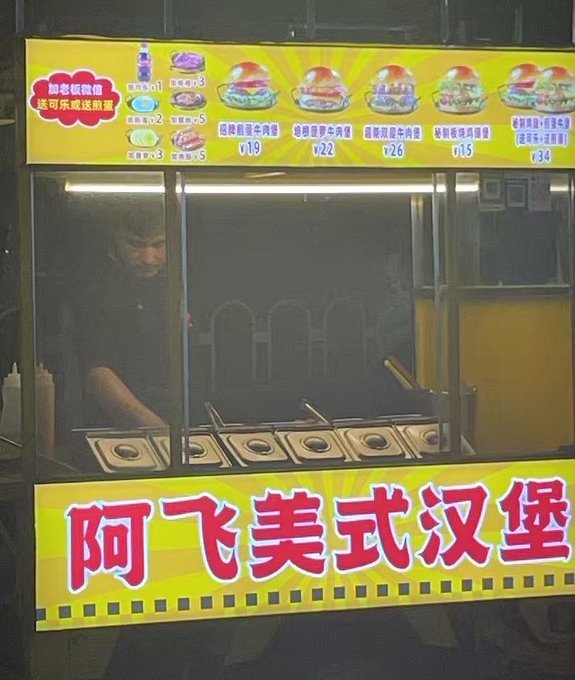A food cart selling burgers with yellow signage reading "阿飞美式汉堡" (Afei American Burger) in Chinese, displaying various burger options priced at ¥19-¥34, with a worker visible inside.
