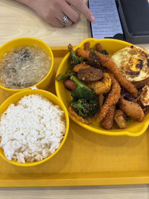 A yellow meal tray holds three bowls: one with plain white rice, one with a clear broth containing vermicelli noodles, and a larger bowl featuring a mixed dish of fried shrimp, broccoli, sausage, and a fried egg. The caption "午饭 2u" (lunch for 2 people) suggests this is a shared meal setup, likely from a cafeteria or food stall where the customer chose a combo meal with rice, soup, and a main dish.