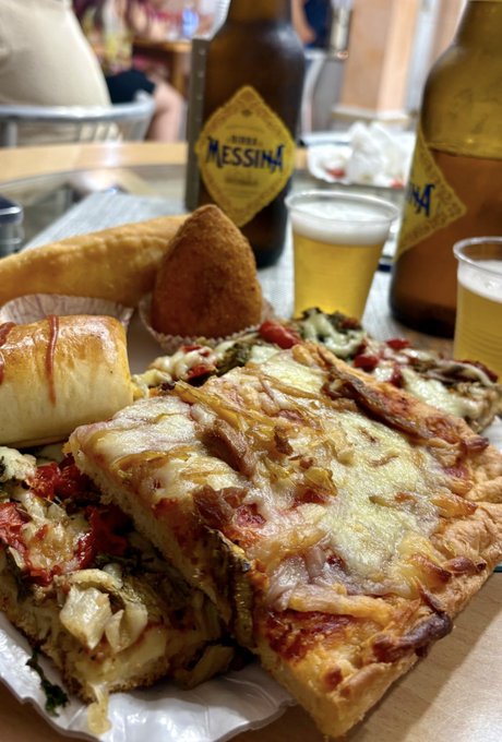 A hearty spread of Sicilian-style pizza, arancini, and a pastry is paired with Messina beer bottles, showcasing a delicious Italian meal that perfectly answers the call for "the best food on earth."