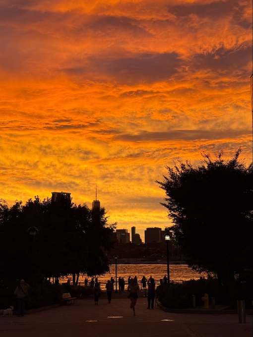 A dramatic orange and yellow sunset sky over a riverside walkway with silhouetted trees, people, and a city skyline in the background.