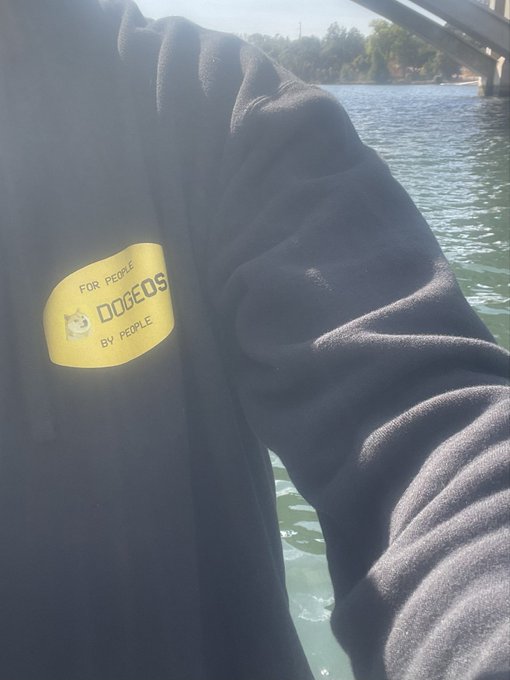A black jacket with a yellow 'FOR PEOPLE, DOGEOS BY PEOPLE' logo patch, photographed outdoors with water and a bridge in the background.