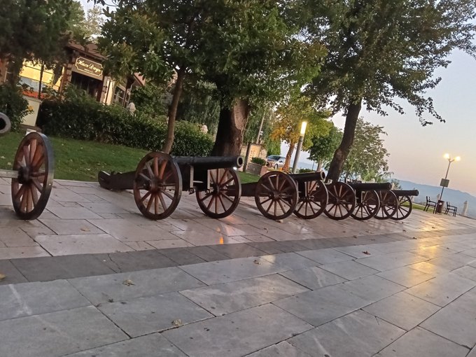 A row of historic wooden cannons on wheeled carriages lines a paved walkway in a serene park setting at dusk, with a partially visible sign reading "...AĞAOĞLU" in the background. The tranquil evening scene complements the post's contemplative message about finding peace, slowing down, and enjoying a quiet good evening, while the Bitcoin hashtag hints at an underlying financial hope or optimism woven into the reflective mood.