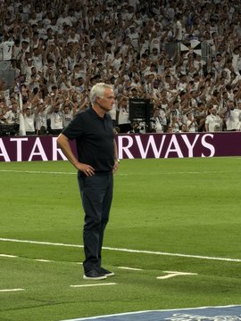 A man with white hair standing on a soccer field with his hands on his hips, with a stadium crowd in the background and "QATAR AIRWAYS" text visible.