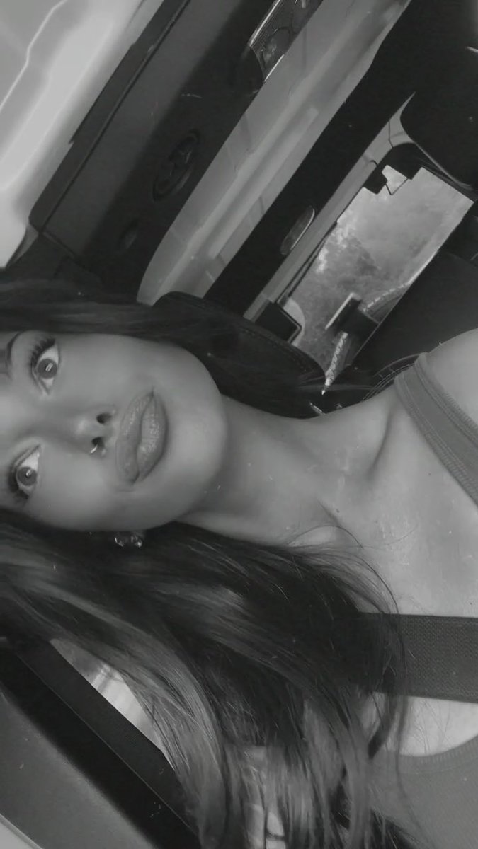 Black and white selfie of a woman with long hair wearing lipstick, taken at a tilted angle inside what appears to be a vehicle.