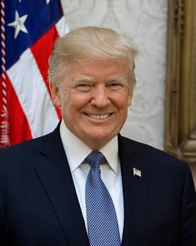 President Donald Trump smiles broadly in a formal portrait, wearing a navy blue suit with a blue patterned tie and an American flag pin, standing in front of a U.S. flag and ornate white architectural details. The image accompanies the post announcing that President Trump has agreed to a new ethics rule in the Clarity Act, with senators set to vote on the final draft tomorrow.