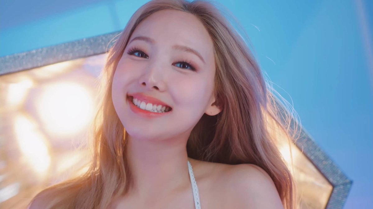A young woman with long blonde hair smiles brightly at the camera, with warm studio lighting and a blue background behind her. The image fits the "Mina next please" caption, likely depicting a K-pop idol being wished for as the next member to appear.