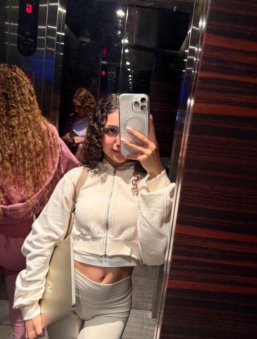 A woman with curly hair takes a mirror selfie in an elevator using a white iPhone, wearing a white zip-up cropped jacket and light gray pants.