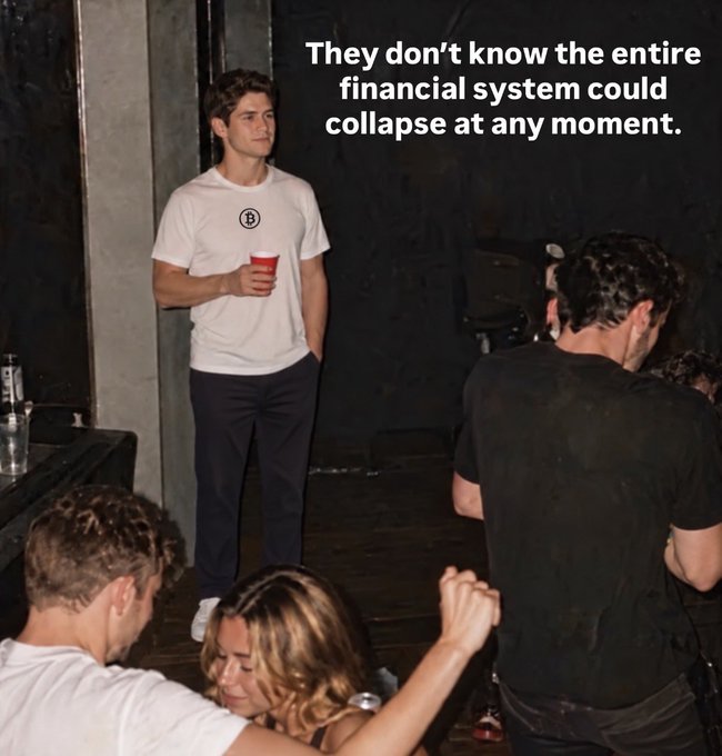 A dim backstage or event setting shows people gathered near a dark wall, while large overlaid text reads, “They don’t know the entire financial system could collapse at any moment.”