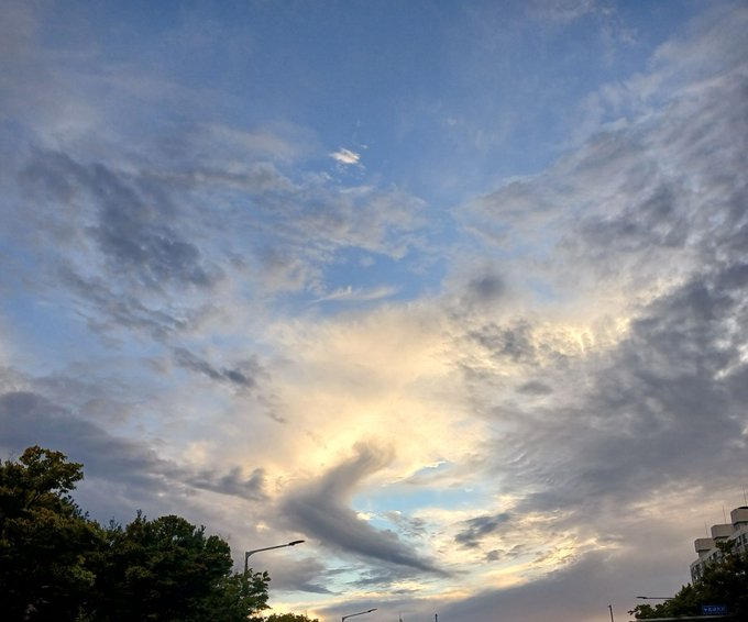 A dramatic sky photograph featuring swirling cloud formations with golden sunlight breaking through, with trees and street lamps visible at the bottom of the frame.