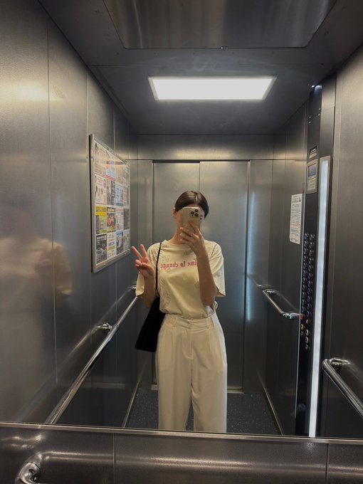 A person taking a mirror selfie inside an elevator, wearing a white t-shirt with red text, white pants, and glasses, while holding a phone and making a peace sign.